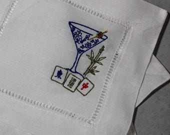 May include: A white square linen napkin featuring an embroidered design of a blue martini glass, a green bamboo stalk, and three mahjong tiles. The napkin has a decorative hemstitched border. The mahjong tiles display blue and red characters.