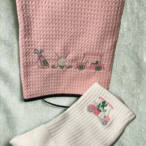 May include: A pink golf towel with a black border and a white golf sock. The towel has an embroidered design of golf clubs, a golf ball, and a golf cart. The sock features a golf cart with a cactus.