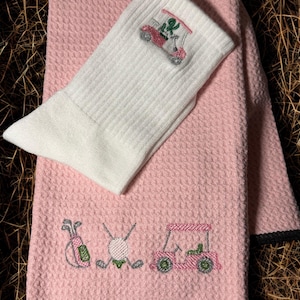 May include: Pink golf-themed towel and sock set. The pink towel has embroidered golf bag, ball, clubs, and golf cart designs. A white sock with a golf cart design is on top of the towel. The towel has a black border.