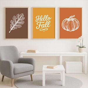 Pode incluir: Três impressões de arte emolduradas com temas de outono. As impressões apresentam uma folha de carvalho branca, o texto "Hello Fall" e uma abóbora, tudo sobre fundos marrons, amarelos e laranja. As impressões são exibidas acima de uma mesa branca e uma poltrona cinza.
