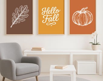 May include: Three framed art prints with autumn themes. The prints feature a white oak leaf, the text "Hello Fall", and a pumpkin, all against brown, yellow, and orange backgrounds. The prints are displayed above a white table and gray armchair.