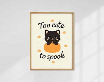 Op de afbeelding: Een ingelijste print met een lichtbeige achtergrond. De print toont een zwarte kat die op een oranje pompoen zit, omringd door kleine pompoenen. De tekst luidt "Too cute to spook" in zwart cursief.