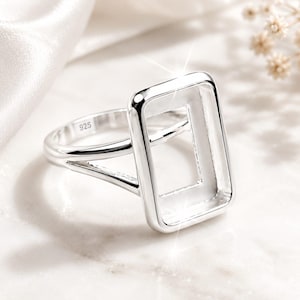 Peut inclure: Bague en argent avec un design rectangulaire ouvert. La bague est en métal poli et présente un anneau divisé. La bague est posée sur un fond blanc doux, avec un subtil effet scintillant.