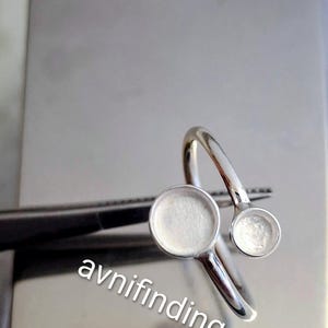 May include: A silver-toned ring with two circular settings. The ring is held by tweezers against a light gray background. The text "avnifinding" is diagonally across the bottom of the image.
