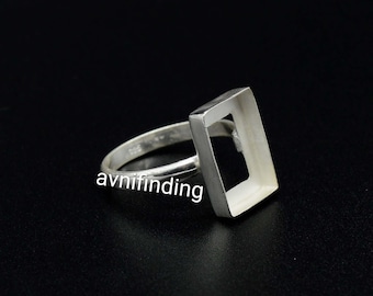 May include: A silver ring setting featuring a rectangular bezel cup designed for stone mounting. The metal band is smooth and polished, with the text avnifinding visible on the surface. This jewellery component provides a frame for custom settings.