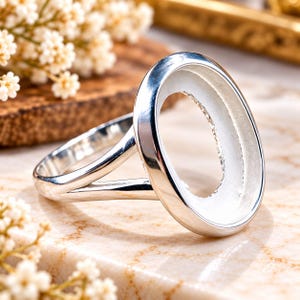 Könnte beinhalten: Ein polierter silberfarbener Ringrohling mit einer glatten, ovalen Fassung für einen Cabochon oder Edelstein. Die Ringschiene ist als geteilte Schiene gestaltet, die an den Seiten des zentralen ovalen Rahmens ansetzt.