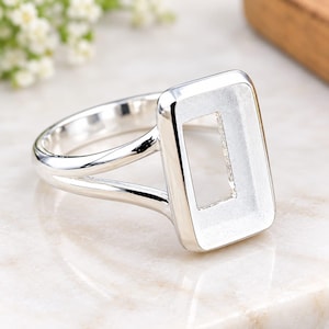 May include: A polished silver-tone ring featuring a rectangular bezel setting with a hollow center. The band is split into two thin, smooth arms that connect to the sides of the rectangular frame, creating a clean and open design for jewelry.