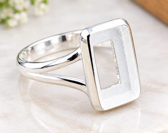 May include: A polished silver-tone ring featuring a rectangular bezel setting with a hollow centre. The band is split into two thin, smooth arms that connect to the sides of the rectangular frame, creating a clean and open design for jewellery.