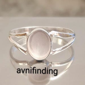 May include: A silver ring featuring a pale pink oval gemstone. The ring has a split band design and the text "avnifinding" is visible at the bottom. The ring is displayed on a reflective surface.