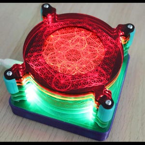 May include: A glowing, geometric LED light. The device features a red, circular top with a complex pattern, supported by teal and green layers. The base is a square shape with a purple border. A white cord extends from the device.