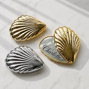 May include: Three shell-shaped compact mirrors with metallic finishes. Two are closed, one in gold and one in silver. The third is open, revealing a mirror with the name Charlotte engraved in black script on the reflective surface of the gold shell.