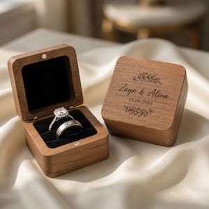 May include: Two square wooden ring boxes with a dark brown finish. One box is open, revealing a silver engagement ring with a square stone and a plain silver wedding band inside on a black velvet insert. The other box is closed with the text Zayn & Alina 12.06.2024 engraved on the lid, framed by two decorative leaf branches.