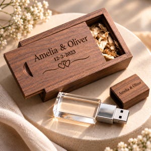 May include: A rectangular dark wood sliding box engraved with the text Amelia & Oliver, the date 12-2-2023, and two hearts. Inside the box are dried flower petals. Beside it lies a clear glass USB flash drive with a matching wood cap engraved with Amelia & Oliver.