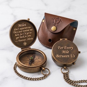 Pode incluir: Duas bússolas de latão em estilo antigo com correntes e uma bolsa de couro castanho. Uma bússola está aberta, mostrando um mostrador e o texto: Miles don't separate us. They just remind me how far I'd go to get back to you. — Forever, Olivia. A outra está fechada, gravada com: For Every Mile Between Us.