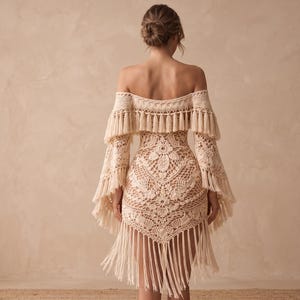 May include: A cream-colored, off-the-shoulder crochet dress with long sleeves and fringe detailing. The dress features a floral pattern on the bodice and a fringed hemline. The sleeves have a bell shape and are adorned with fringe.
