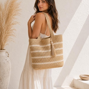 May include: A woven straw tote bag featuring horizontal white stripes against a natural tan background. The bag includes two shoulder straps and a decorative tassel accessory with a small wooden bead attached to one of the handles.