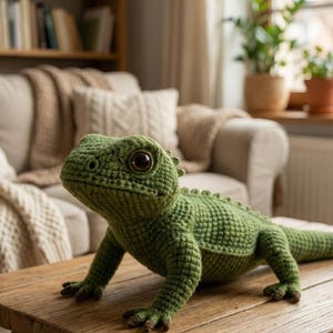 Peut inclure: Un lézard vert crocheté à la main, avec des écailles détaillées et de grands yeux foncés. Le reptile en peluche est posé sur une table en bois, mettant en valeur ses pattes et sa queue. Un article artisanal unique.