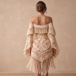 May include: A cream-colored, off-the-shoulder crochet dress with long sleeves and fringe detailing. The dress features a floral pattern on the bodice and a fringed hemline. The sleeves have a bell shape and are adorned with fringe.