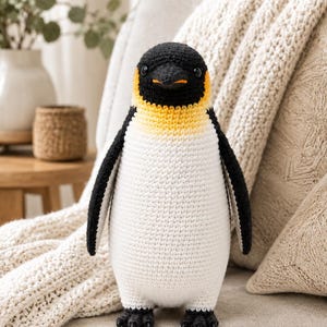 Op de afbeelding: Een gehaakt pinguïn knuffeltje met een zwarte kop en rug, een witte buik en een geel-oranje kleurverloop rond de nek. De pinguïn heeft kleine zwarte oogjes, een puntige zwarte snavel en zwarte voetjes, gemaakt met zichtbare garensteken.