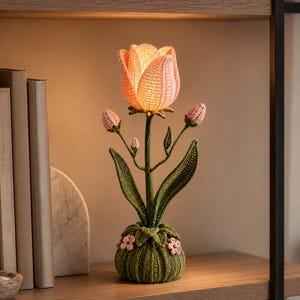 Puede incluir: Lámpara de tulipán hecha a mano con ganchillo, con una flor rosa suave y tallo y hojas verdes. La lámpara tiene una base redonda verde con detalles de flores rosas. La lámpara está encendida, emitiendo un brillo cálido.