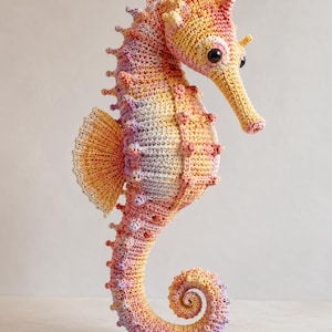 May include: A crocheted seahorse figurine featuring a gradient of yellow, orange, pink, and light purple yarn. The creature has a textured body with small protruding spikes, a fan-shaped dorsal fin, and large, dark, glossy plastic safety eyes.