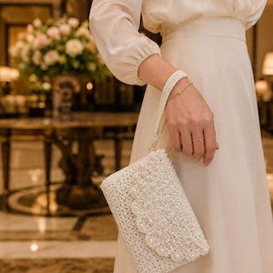 Könnte beinhalten: Elfenbeinfarbene Häkeltasche mit einem wellenförmigen Klappverschluss, verziert mit kleinen Perlen. Die Tasche hat eine Handschlaufe und eine goldfarbene Kette. Das Design ist elegant und für besondere Anlässe geeignet.