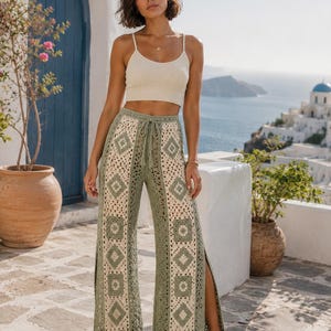 Puede incluir: Una mujer con un top corto color crema y pantalones de crochet verde oliva con un patrón geométrico. Los pantalones tienen cintura con cordón y aberturas laterales. El fondo muestra un paisaje costero con edificios blancos y una puerta azul.