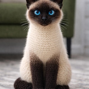 以下が含まれることがあります： 鮮やかな青い目とクリーム色の体をした、かぎ針編みのシャム猫。猫の顔、耳、足、尾は濃い茶色です。模様のあるラグの上に座っており、手作りのアイテムの細部を示しています。