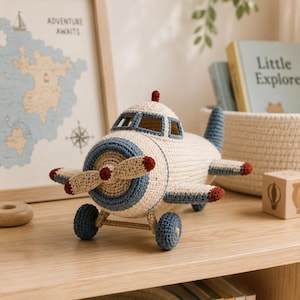 Può includere: Un aereo giocattolo all'uncinetto color crema, blu e rosso. L'elica, le ali e le ruote sono dettagliate. Il giocattolo è su una mensola di legno, con una mappa incorniciata e libri per bambini sullo sfondo.