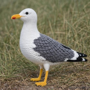Op de afbeelding: Een gehaakte zeemeeuw met een wit lichaam, grijze vleugels en zwarte vleugeltips. De zeemeeuw heeft een gele snavel met een rode punt en gele poten. De vogel staat op de grond met gras op de achtergrond.