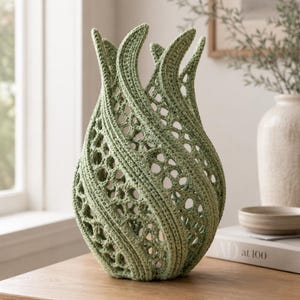 May include: A sage green crochet vase with a sculptural, flame-like silhouette. The object features a dense, solid vertical stitch pattern alternating with open, web-like lattice sections that create a hollow, airy structure throughout the body.