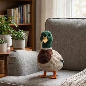 May include: A handmade crochet duck with a green head, brown chest, and beige body. The duck has orange feet and a yellow beak. It is sitting on a gray armchair in a cozy living room setting.