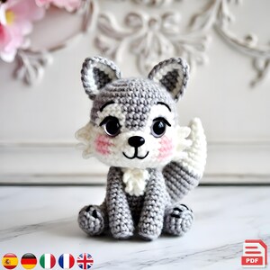 May include: A crocheted gray and white wolf figurine with large black eyes, pink cheeks, and a white chest. The handmade stuffed animal is sitting upright with its tail curled to the side. The wolf has a cute, friendly expression.