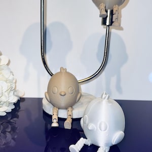 Könnte beinhalten: Eine dekorative Präsentation mit mehreren vogelförmigen Figuren in Weiß und Beige. Die Vögel haben bewegliche Beine und sitzen auf einem Metallständer und einer Marmorbasis. Der Hintergrund ist eine dunkelblaue Oberfläche.