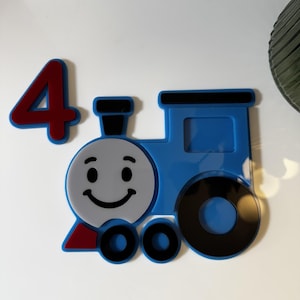 May include: A blue train with a smiling face, black wheels, and a red triangle. A red number 4 is next to the train. The train is a decorative item.