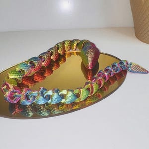 May include: A multi-coloured, articulated snake toy in shades of red, blue, green, and yellow, on a gold-coloured oval tray. The snake's body is segmented, allowing for flexible posing. The tray reflects the snake's image.