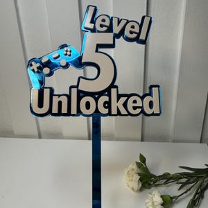 May include: A blue and white cake topper with the words "Level 5 Unlocked" and a controller graphic. The number 5 is in the center, and the topper is held by a blue stem. White flowers are in the background.
