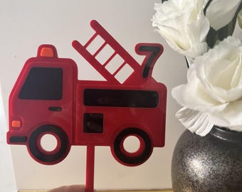 May include: A red fire truck cake topper with black accents, a ladder, and the number 7. The topper is held in front of a vase of white flowers.