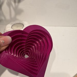 Könnte beinhalten: Ein magenta-farbenes, herzförmiges Dekorationsobjekt mit einem geschichteten Design. Das Herz hat mehrere verschachtelte Schichten, die einen dreidimensionalen Effekt erzeugen. Das Objekt ist wahrscheinlich aus Holz oder einem ähnlichen Material.