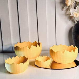 Peut inclure: Quatre bols décoratifs jaunes avec un bord dentelé, ressemblant à des coquilles d'œufs, sont disposés sur une surface réfléchissante. Les bols varient en taille, le plus grand étant à l'arrière et le plus petit à l'avant. Un vase de fleurs est en arrière-plan.