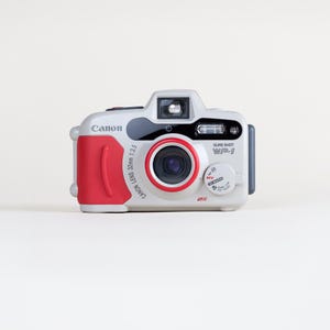 Puede incluir: Una cámara de película Canon Sure Shot WP-1 de color gris claro con empuñadura roja y anillo de lente rojo. La parte frontal presenta un objetivo de 32mm 1:3.5, un flash y un dial circular con ajustes para apagado, automático y un enfoque de 0,45m a infinito.