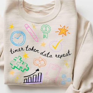May include: A sand-coloured sweatshirt featuring colourful doodle graphics, such as a clock, pen, star, checkmark, puzzle pieces, and a graph. The text "timer token data repeat" is written in black script.