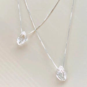 Op de afbeelding: Twee zilveren kettingen met heldere, onregelmatig gevormde kristallen hangers. De hangers hangen aan fijne zilveren kettingen. De kettingen worden gepresenteerd tegen een lichte achtergrond, waardoor de helderheid van het kristal en de glans van het zilver worden benadrukt.