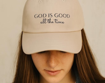 May include: A cream-coloured baseball cap featuring the words "GOD IS GOOD all the time" in black. The cap is worn by a person wearing a denim jacket and a white t-shirt.