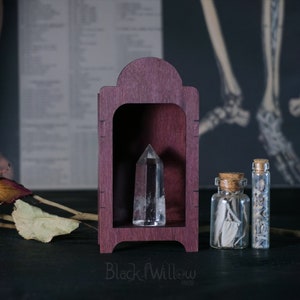 Puede incluir: Una pequeña vitrina de madera con un cristal de cuarzo transparente en su interior. La vitrina es de color burdeos oscuro y tiene un arco decorativo en la parte superior. Dos pequeños frascos de vidrio con tapas de corcho están sobre la mesa junto a la vitrina. Un frasco contiene hierbas secas y el otro contiene salvia blanca.