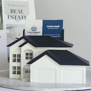 May include: A white miniature house with a black roof, designed to hold business cards. The house has a two-car garage and multiple windows. Business cards with the text "REAL ESTATE" and "YOUR NAME" are displayed in the house.