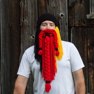 May include: A person wearing a black knit hat with a long, braided yarn beard in black, red, and yellow.