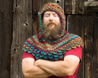 May include: A person wearing a multi-colored crocheted hooded cowl with a scale pattern. The cowl is made with a variety of shades of green, blue, purple, and brown yarn. The person is wearing a red shirt and has a beard.