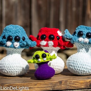 May include: Four crocheted mushroom plushies with googly eyes. The mushrooms are blue, red, green, and purple. They are sitting on a wooden surface.