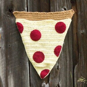 Puede incluir: Una manta de crochet en forma de rebanada de pizza con una corteza marr&oacute;n y una base de queso blanco. La manta tiene c&iacute;rculos de pepperoni rojos.